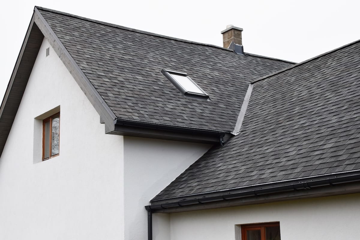 Part of white house with new wooden eaves, plastic rain gutter, window and chimney in grey shingles roof and brown window in facade. best roof shingles