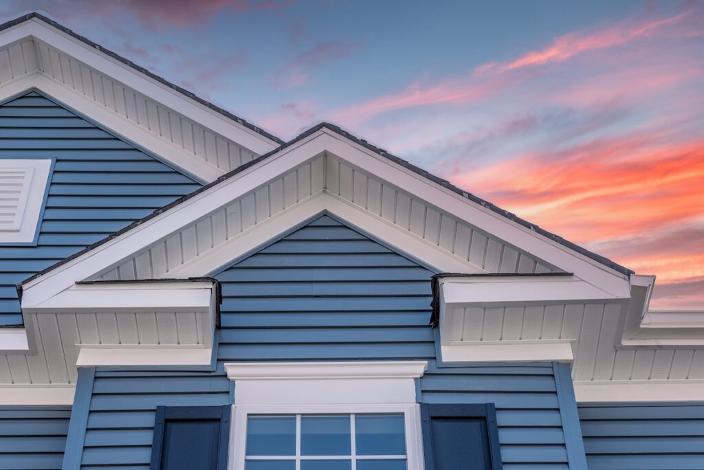 soffit and fascia repair Triangle shape decorative gable with colonial white soffit and fascia on a blue horizontal vinyl siding modern American estate home with colorful dramatic stunning orange sunset sky