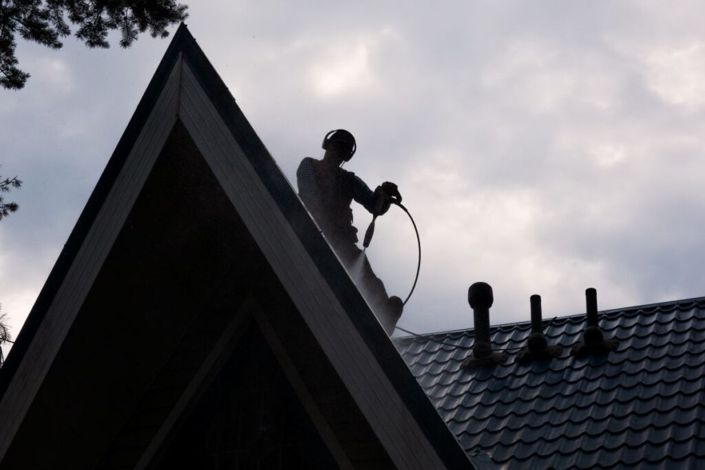 A man sits on a gabled roof made of metal tiles and washes the surface