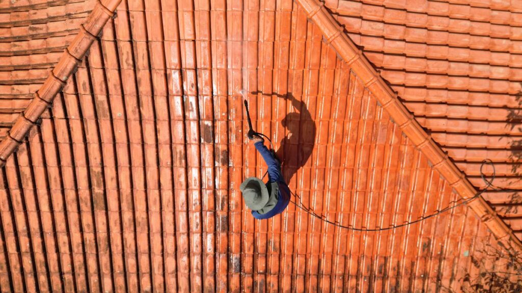 A person outdoors cleaning the red clay roof using a high pressure washer