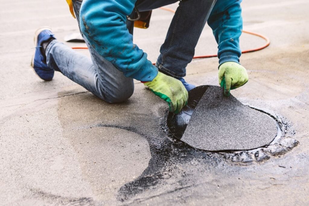 Roofer repairing a damaged section of a roof. Worker kneels on a wet, flat rooftop to apply a circular patch